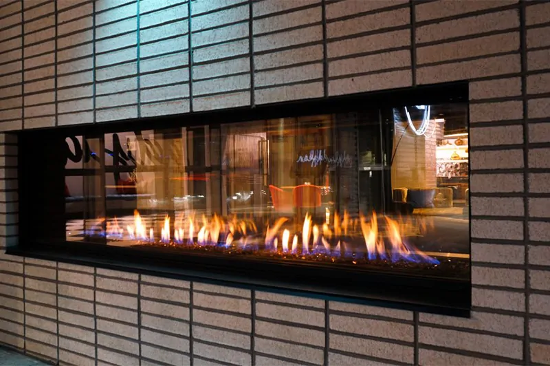 Close-up of a modern linear See Through 100" Flare Fireplace set in a brick wall with a cozy indoor space visible through the glass.