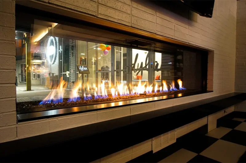 Flare See Through 100" Modern Commercial Fireplace with 24" High Cool Touch Double Glass; featuring a white brick surround and a view of a modern storefront.