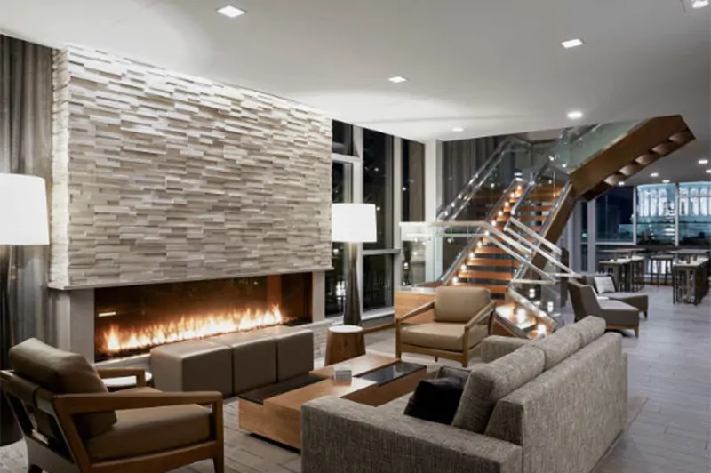 Modern lounge with a linear See Through 100" Modern Commercial Fireplace with 24" High Cool Touch Double Glass, stacked stone surround, and glass staircase.